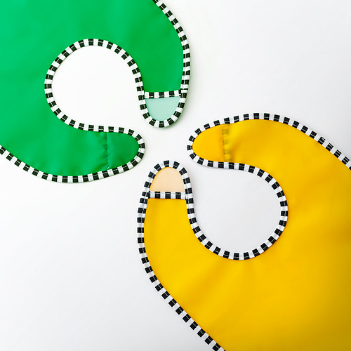 Tres baberos de colores brillantes, verde, beige y amarillo, colocados planos, bordeados con ribetes a cuadros blancos y negros.