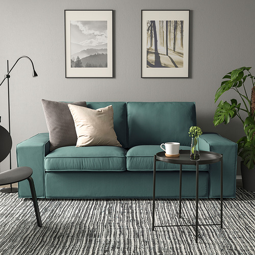 Acogedora escena de sala de estar con sofá KIVIK en verde, almohadas de felpa, pequeña mesa de metal, plantas, taza y láminas de arte en paredes grises.
