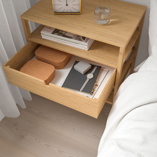 Mesita de noche con cajón abierto que muestra revistas y tacos de corcho. Reloj y cristal en la parte superior, ropa de cama blanca cerca.