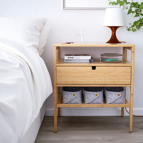 Mesita de noche pequeña de bambú claro con un cajón y estantes abiertos. Cabe perfectamente al lado de una cama, sosteniendo una lámpara, libros y contenedores de almacenamiento.