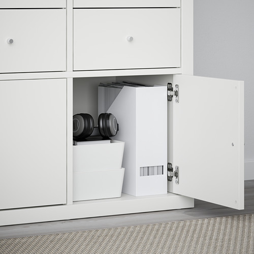 Armario blanco con puerta abierta que muestra mancuernas negras y cajas de almacenamiento. Forma parte de la serie KALLAX.