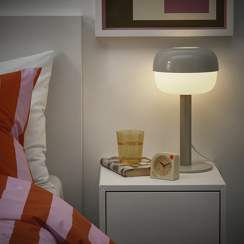 Acogedor dormitorio con una mesita de noche moderna, lámpara gris, reloj amarillo, vaso de agua y libro doblado, creando un ambiente tranquilo.
