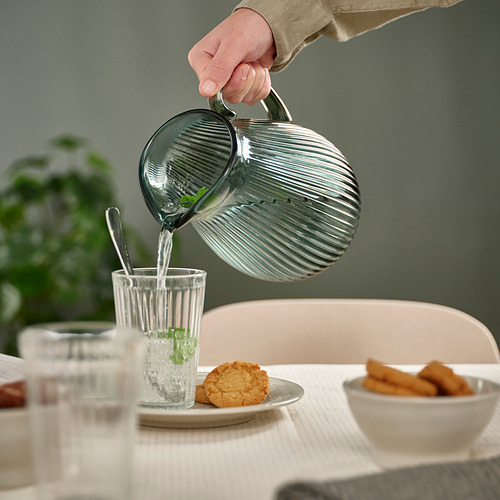 La mano vierte agua de una jarra de vidrio acanalado en un vaso alto junto a platos con galletas y bocadillos fritos en una mesa.