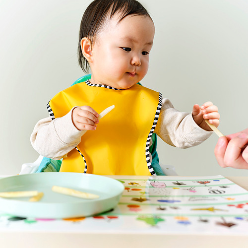Bebé con babero amarillo, sostiene la cuchara, la mano del adulto ofrece otra. La mesa tiene notas adhesivas de colores.