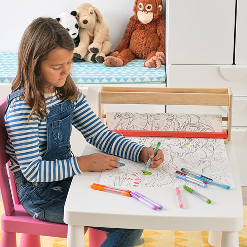 Una niña se sienta en una mesa para niños, dibujando en un rollo de papel con marcadores. La mesa contiene bolígrafos y materiales de arte, fomentando la creatividad.