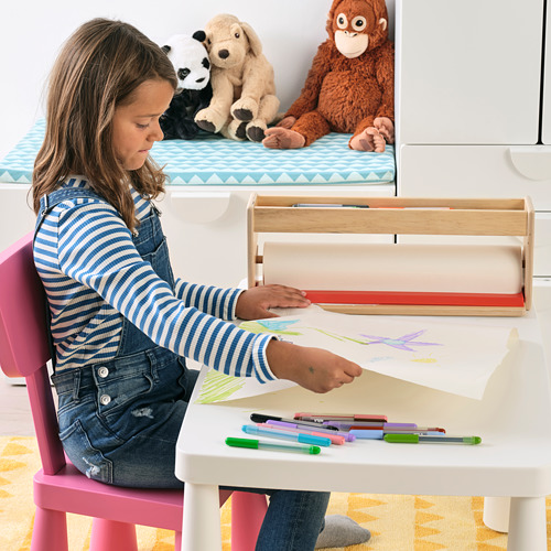 Una niña está sentada en el escritorio de un niño, dibujando en un rollo de papel. El soporte organiza bolígrafos y lápices de colores. Los animales de peluche son visibles en un estante detrás de ella.