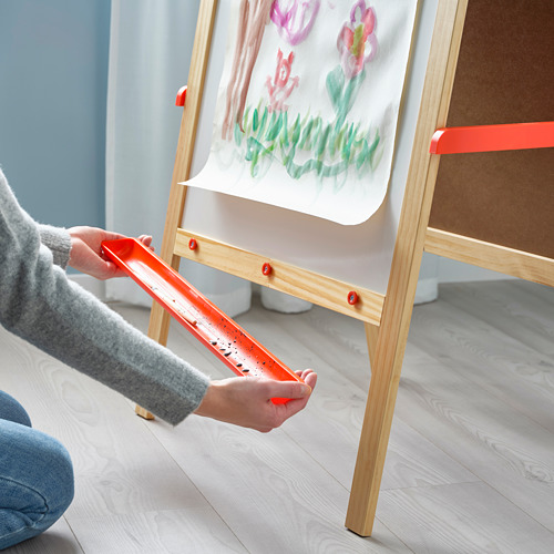 Manos de un niño sosteniendo unas tijeras de juguete naranjas, recortando papel en un caballete de madera con un dibujo de flores.