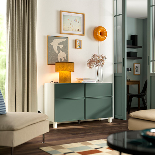 Habitación moderna con gabinete BESTÅ en color verde azulado y blanco, lámpara, obras de arte y cortinas neutras, adyacente a una elegante puerta de vidrio.