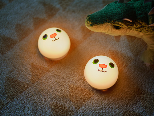 Tres luces nocturnas redondas y blancas con caras de dibujos animados en un resplandor suave y cálido, junto a un peluche de dinosaurio verde sobre un suelo alfombrado.