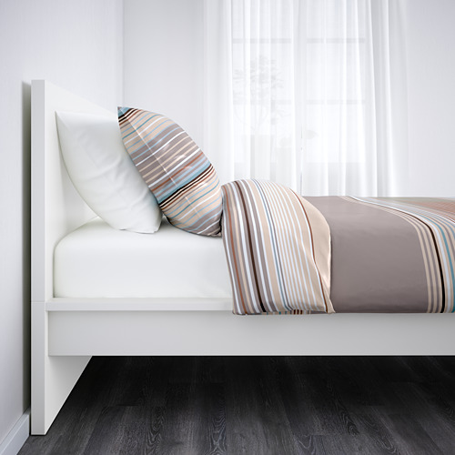 Cama minimalista con almohadas y manta de rayas, cabecero alto contra pared blanca, con suelo de madera oscura.
