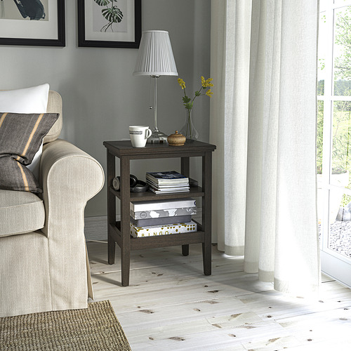 Mesa auxiliar de IDANÄS junto a silla gris, sosteniendo lámpara, tazas, libros. Madera oscura, suelos blancos. Ideal para espacios reducidos.