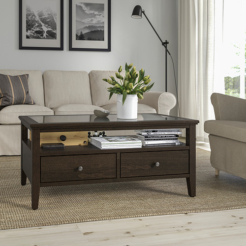 Mesa de centro IDANÄS con tapa de cristal, central en la sala de estar, rodeada de sofá y sillón beige, con alfombra, jarrón, libros, lámpara y arte.