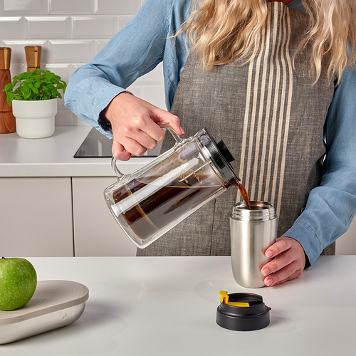 Una persona vierte café de una jarra transparente en una taza de viaje ENVÄLDIG de acero inoxidable con tapa negra sobre la encimera de la cocina.