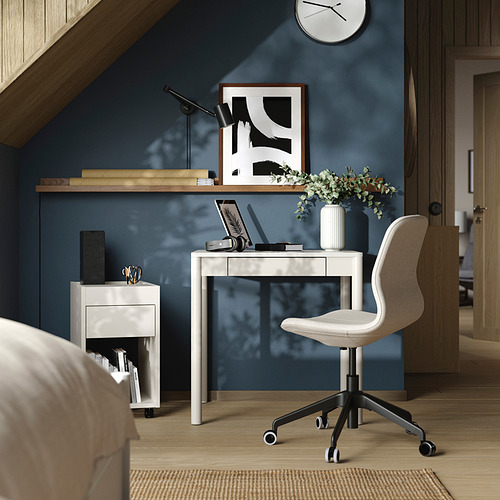 Moderna oficina en casa con escritorio TONSTAD blanco, silla y pared azul oscuro. El escritorio tiene una computadora portátil, flores y un reloj.