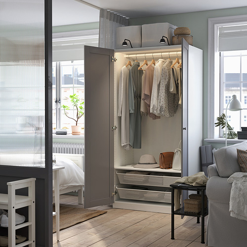 Moderno armario abierto con ropa, sombreros y bolsos, estantes superiores iluminados, sofá gris adyacente y cama blanca, paredes grises.