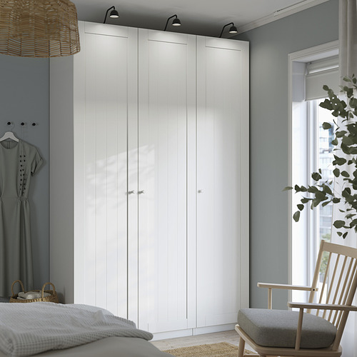 Moderno armario blanco PAX en el dormitorio, alto con líneas elegantes. Silla de madera, ropa y una gran ventana al lado.