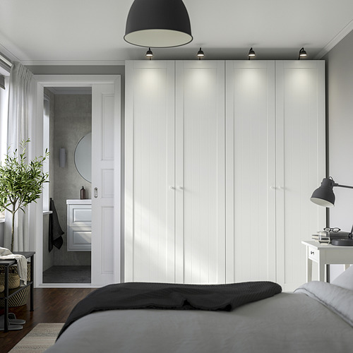 Dormitorio moderno con amplio closet blanco PAX, cama gris, planta y lámpara de escritorio. La iluminación tenue resalta los armarios de diseño alto y elegante.