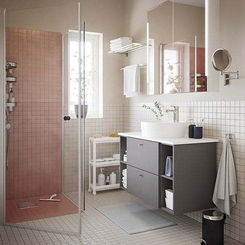 Baño moderno con ducha de vidrio, lavabo blanco, gabinete gris y carrito de estante blanco VESKEN debajo de la ventana.