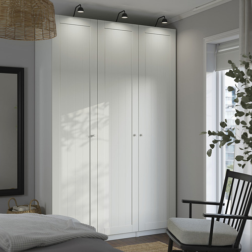 Dormitorio moderno y minimalista con un gran armario blanco PAX, pared alta y amplia, junto a una ventana con marco negro y una silla de madera.