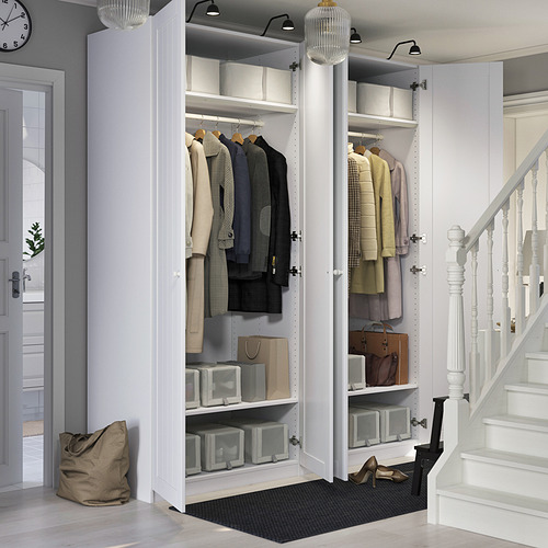 Armario PAX blanco con puertas abiertas que muestran ropa, estantes y cajas de almacenamiento. Situado cerca de una entrada con escaleras y una puerta.