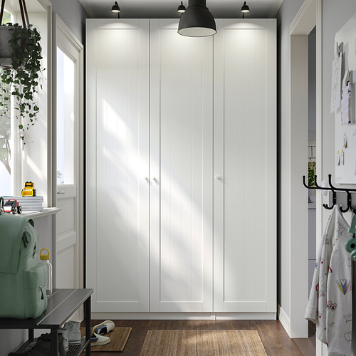 Un armario blanco PAX, alto y elegante, con las puertas cerradas. Es el mueble principal, más grande que los elementos circundantes, con luces colgantes en la parte superior.