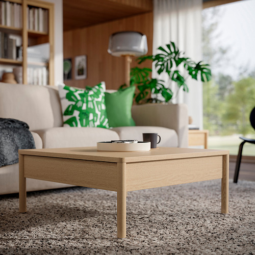 Mesa de centro de chapa de roble marrón con cajones laterales sobre alfombra beige en acogedora sala de estar con sofá, planta y estantería.