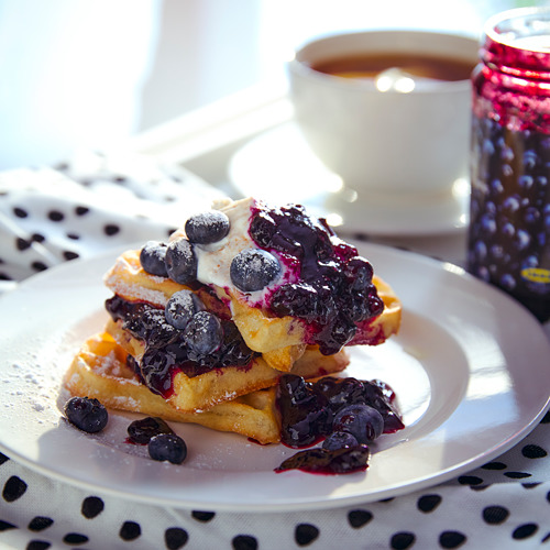 Un plato de waffles cubierto con mermelada de arándanos y arándanos frescos, acompañado de una taza de café.