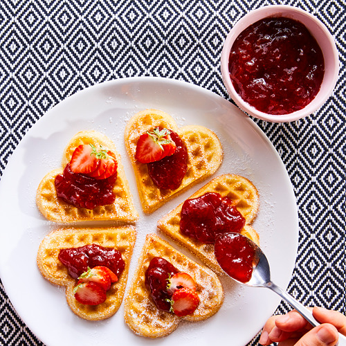 Gofres en forma de corazón con mermelada roja y fresas en un plato blanco. Una cuchara con mermelada al lado.