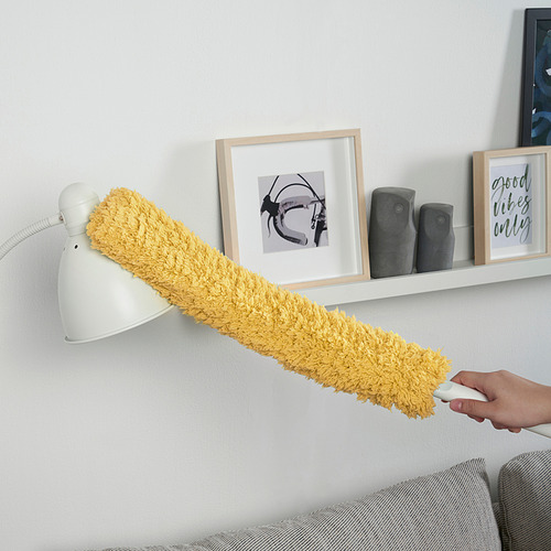 La mano sostiene el plumero de microfibra amarilla, limpiando sobre el sofá. Lámpara de pared blanca y estante con arte enmarcado detrás.