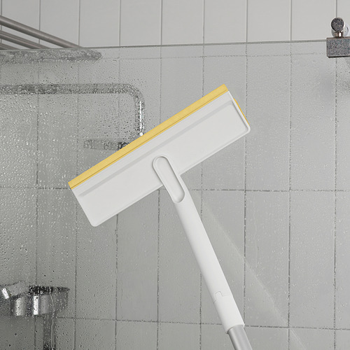 Un mango de limpieza telescópico blanco con un accesorio de escobilla de goma amarilla y blanca, que limpia la pared de la ducha con gotas de agua.