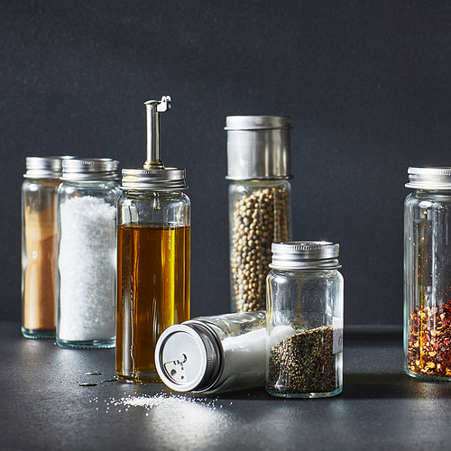 Seis recipientes de vidrio con tapas metálicas y contenido que incluye sal, aceite, especias y hojuelas de chile seco, dispuestos cuidadosamente sobre una superficie oscura.