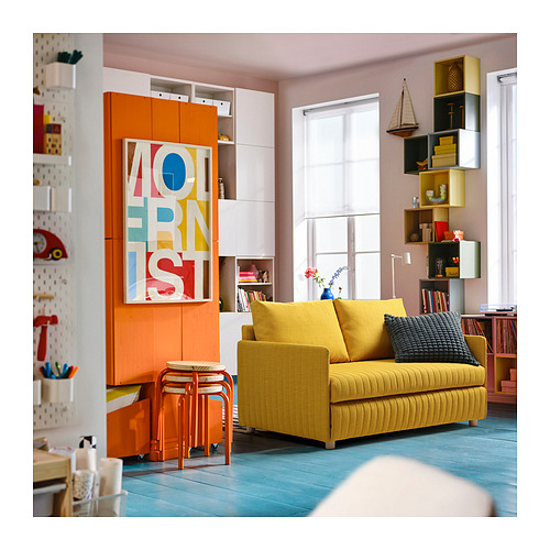 Sala de estar luminosa con sofá amarillo FRIDHULT, arte vibrante y estantes multicolores.