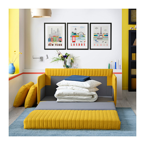 Sala de estar moderna con sofá cama amarillo FRIDHULT, almohadas blancas, jarrón azul, impresiones artísticas de Nueva York, Londres, París, accesorios coloridos.
