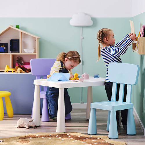 Niños jugando con el juego de té de juguete en una mesa colorida, uno en una silla azul, otro en un taburete morado, una casa de muñecas blanca cerca.