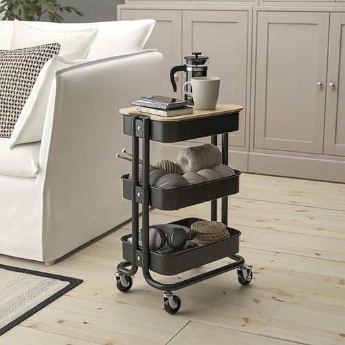 Pequeño carrito RÅSKOG negro cerca del sofá blanco, que se coloca debajo de las mesas. Sostiene cafetera, tazas e hilo. Fabricado en acero y bambú.