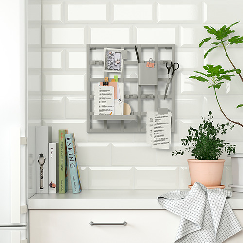 El organizador de cocina en la pared contiene notas, llaves, tijeras, libros y plantas. Fondo de azulejos blancos, tela a cuadros y vegetación en macetas.