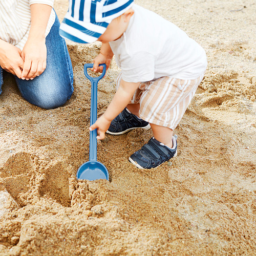 Un niño juega con una pala de arena azul en una superficie arenosa, probablemente en una playa, con un adulto cerca.