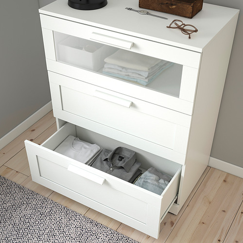 Cómoda blanca con cajón abierto que muestra la ropa doblada, cajón superior transparente, sobre suelo de madera.