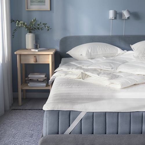 Dormitorio azul con protector de colchón gris, ropa de cama cuidada, mesita de noche con plantas, libros y lámpara.