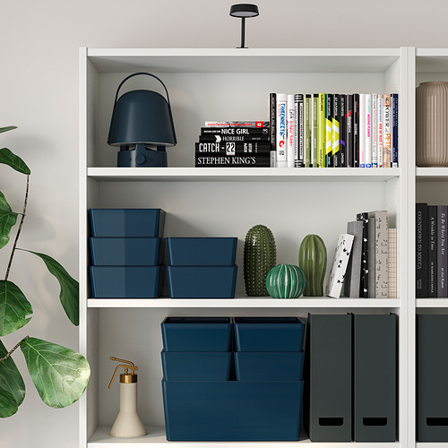 Estantería blanca con cajas de almacenamiento azules, libros, lámpara y planta.