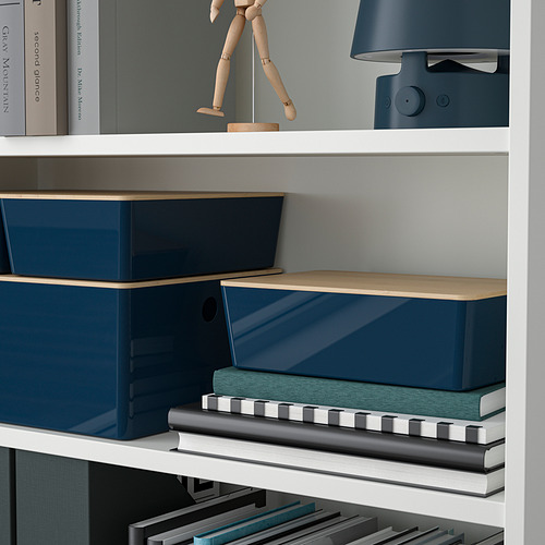 Una estantería blanca con cajas de almacenamiento azules y tapas de madera organiza varios objetos, incluyendo libros, una figura de madera y un altavoz.