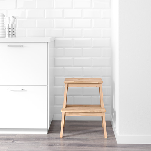 Un taburete de madera con dos escalones en una habitación blanca, colocado contra una pared de ladrillo blanco y junto a armarios blancos.