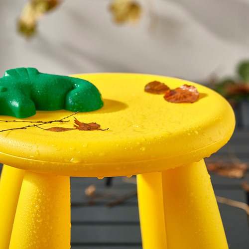 Un taburete infantil amarillo con patas, mojado del exterior, que sostiene hojas y formas de juguete en la parte superior.