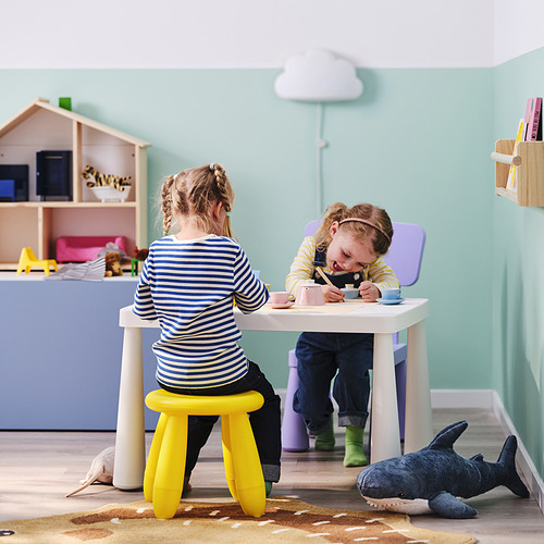 Dos niños jugando con juguetes en una habitación colorida. Un tiburón azul de juguete está sobre la alfombra y una casa de muñecas está contra una pared azul claro.