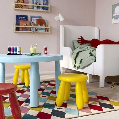Sala de juegos para niños con mesa azul pastel MAMMUT, pequeños taburetes amarillos y rojos, alfombra colorida, cama blanca con ropa de cama, estantes de pared con libros para niños.