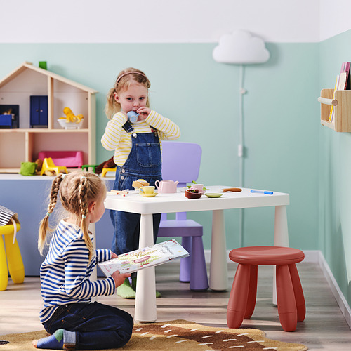 Dos niñas jugando en una habitación colorida. Uno sostiene un libro, el otro bebe de una taza en una pequeña mesa blanca para niños.