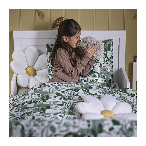 La niña besa el peluche de la cama con sábanas de hojas verdes y almohadas de margaritas de poliéster reciclado.