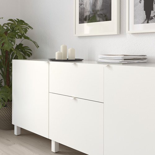 Armarios blancos con manijas minimalistas, velas y libros apilados en la parte superior, contra una pared clara con fotos enmarcadas y una planta verde cerca.