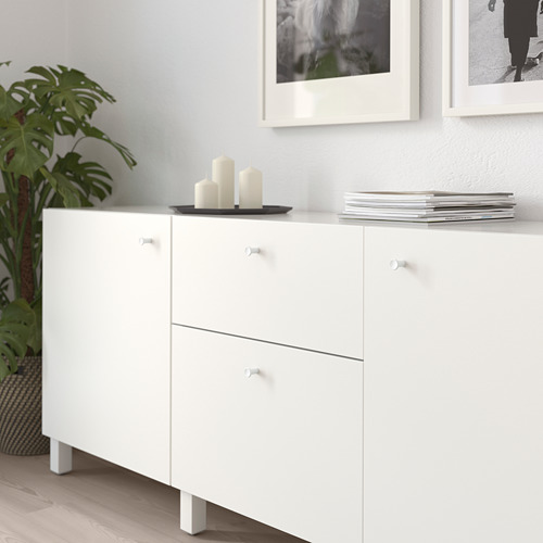 Mueble blanco con cajones y puertas, con varios pomos. En la parte superior hay velas y una pila de revistas. Junto a una planta verde en una cesta.