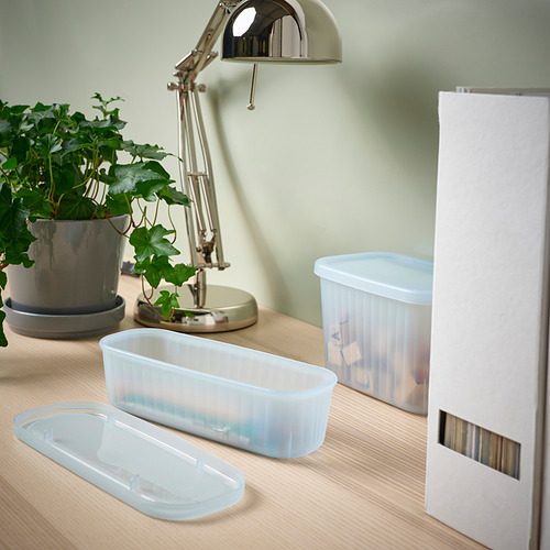 Escritorio con cajas de almacenamiento transparentes RYKTA, lámpara y planta. Las cajas son pequeñas, apilables y parcialmente transparentes.